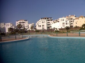 Ein Pool mit gewölbter Kante, umgeben von einem Zaun, mit Gebäuden mit Balkonen im Hintergrund.