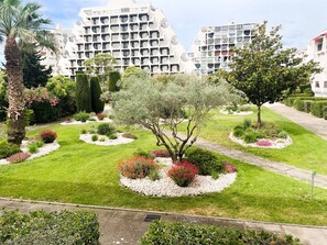 A landscaped garden with a variety of plants and trees, a pathway, and modern buildings in the background. - Air-conditioned apartment in city center - 2 guests (La Grande-Motte)