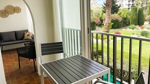 A balcony with a table and a chair, a sofa, and a view of a garden and buildings. - Air-conditioned apartment in city center - 2 guests (La Grande-Motte)