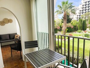A balcony with a table and a chair, a sofa, and a view of a garden and buildings. - Air-conditioned apartment in city center - 2 guests (La Grande-Motte)