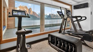 Ein Fitnessraum mit Laufbändern, einem Ergometer und einer Gewichtbank, mit großen Fenstern und Blick auf die Berge.