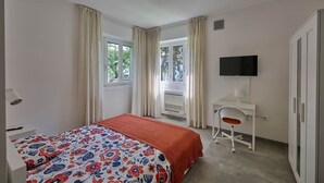 Ein Schlafzimmer mit Bett, Schreibtisch, Stuhl, Fernseher und einem Fenster mit Vorhängen.