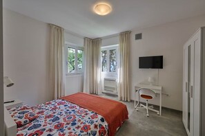 Ein Schlafzimmer mit Bett, Schreibtisch, Stuhl, Fernseher und einem Fenster mit Vorhängen.