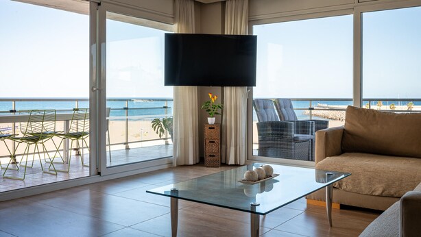 Amplia sala de estar con una gran mesa de vidrio, un sofá y un televisor de pantalla plana. La habitación tiene ventanas grandes que ofrecen vistas a la playa y al océano.