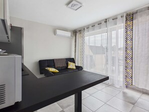 A modern room with a black sofa, a small table, a white air conditioner, and a patterned curtain. - Cozy apartment, south terrace - 4 guests (Marseillan-Plage)
