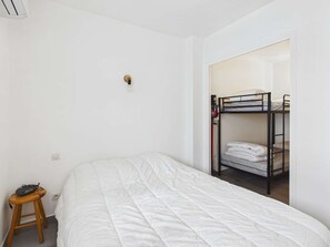 A single bed with a white duvet, a wooden stool, a power outlet, and a bunk bed in the background. - Cozy apartment, south terrace - 4 guests (Marseillan-Plage)