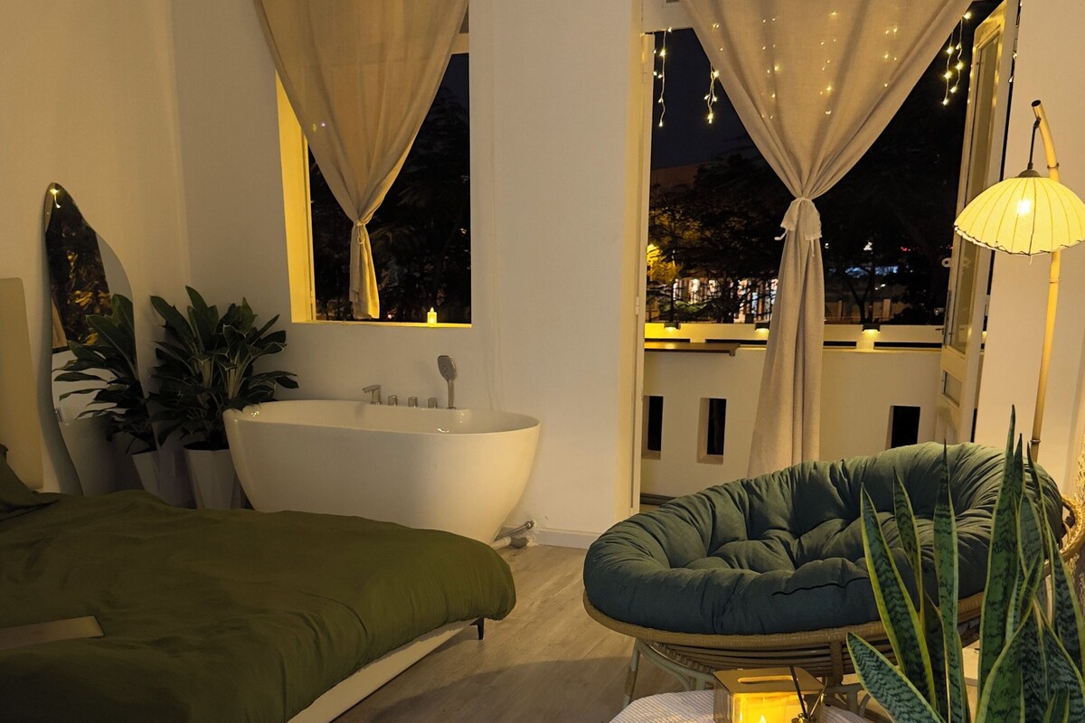 A modern bedroom with a large window, a bathtub, a green sofa, and a potted plant.
