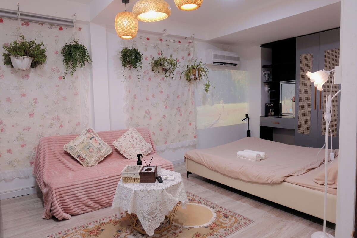 A bedroom with a pink sofa, a bed, a small table with a radio, and hanging plants.