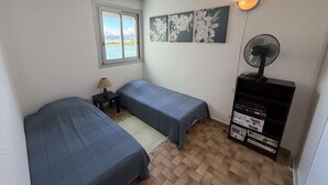 A room with two beds, a window with a view, a wall-mounted fan, and a shelf with books. - Apartment with parking - 4/6 guests (Port Leucate)