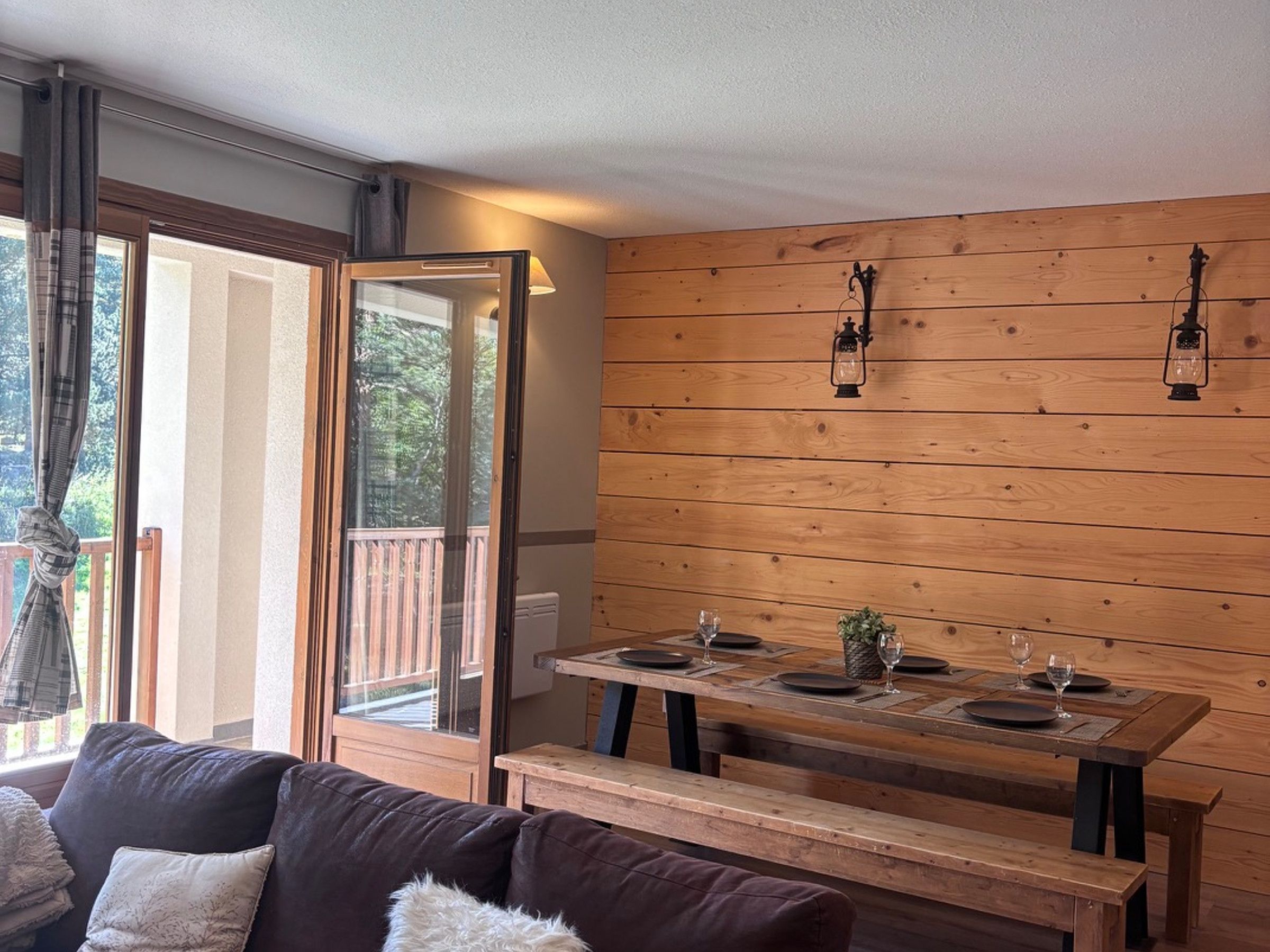 A dining area with a wooden table set for two, a bench, and a view through a glass door to an outdoor deck.