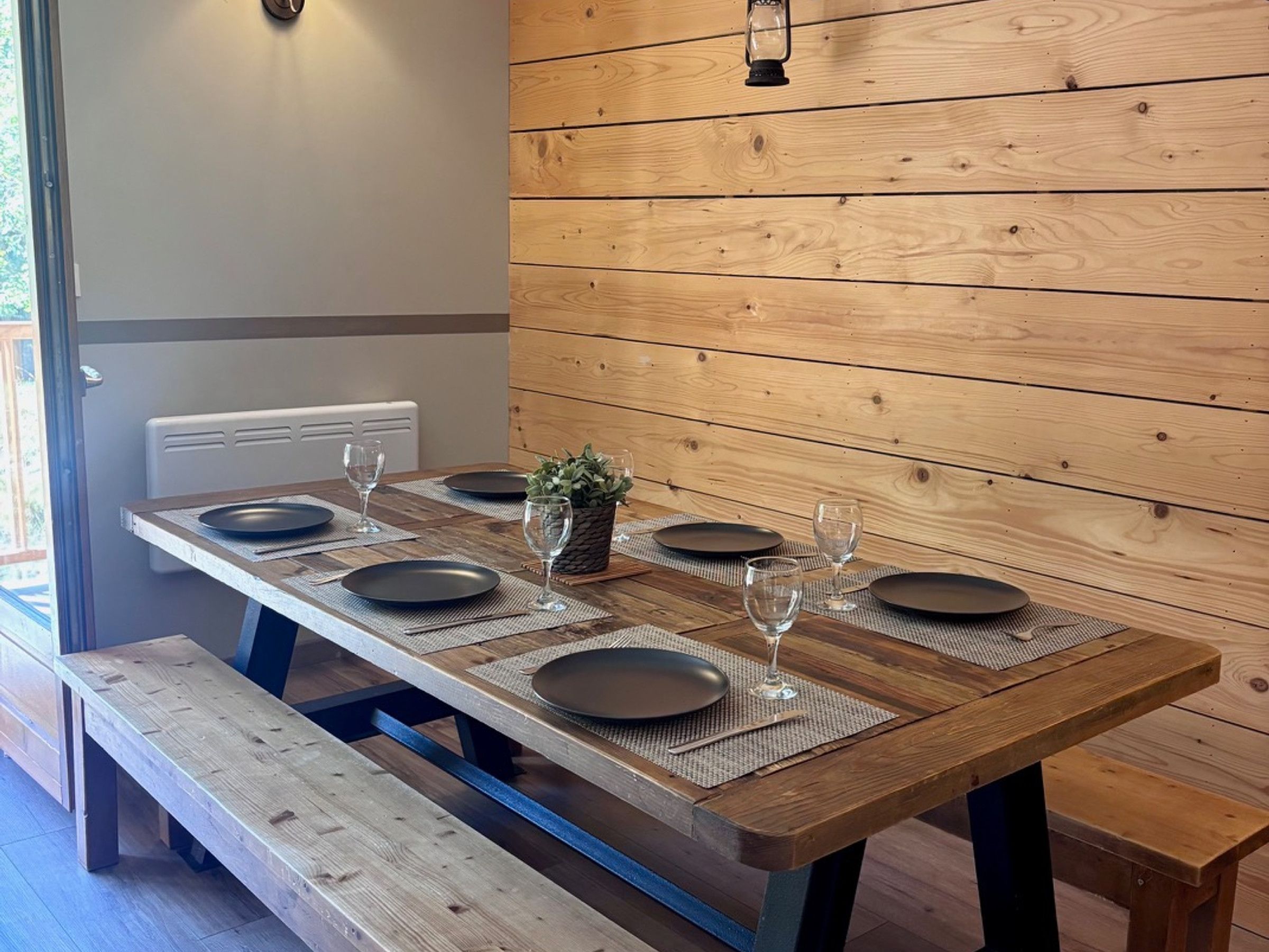 A dining table set with plates, glasses, and a small plant, against a wooden wall with a radiator.
