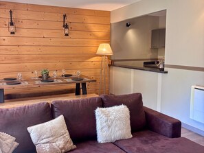 Ein modernes Wohnzimmer mit Holzwand, einem Esstisch für vier Personen, einer Couch mit dekorativen Kissen und einer Stehlampe.