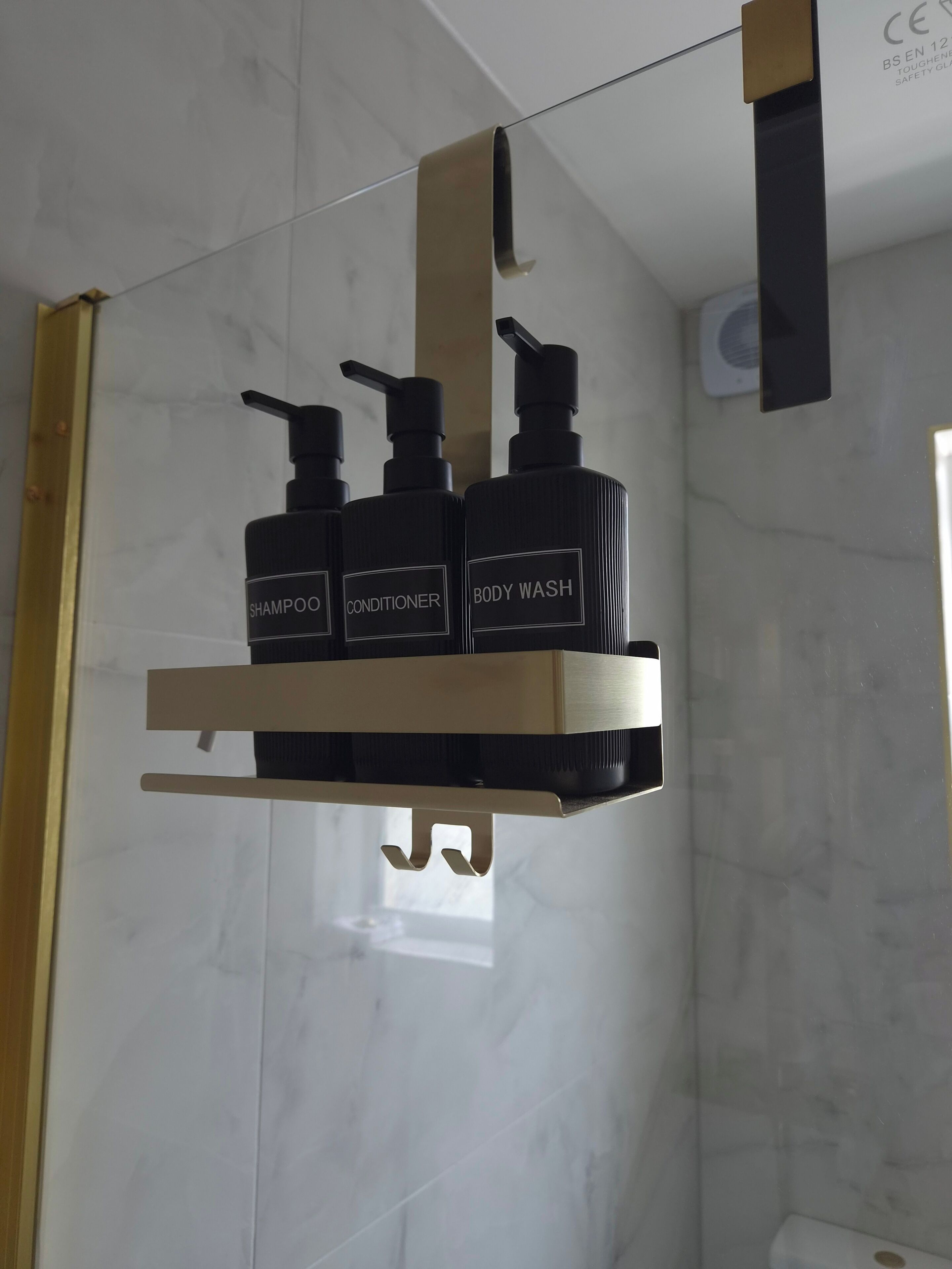 A bathroom with a marble wall, a shower area, and a shelf holding three bottles labeled 'SHAMPOO,' 'CONDITIONER,' and 'BODY WASH.'