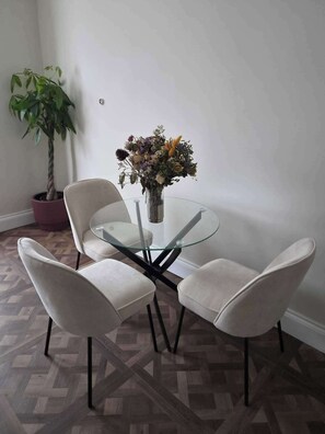 A dining area with a glass table, four white chairs, a potted plant, and a vase with flowers. - Spacious Apartment Sleeps 2 With Parking, Newly Refurbished (Ilford)