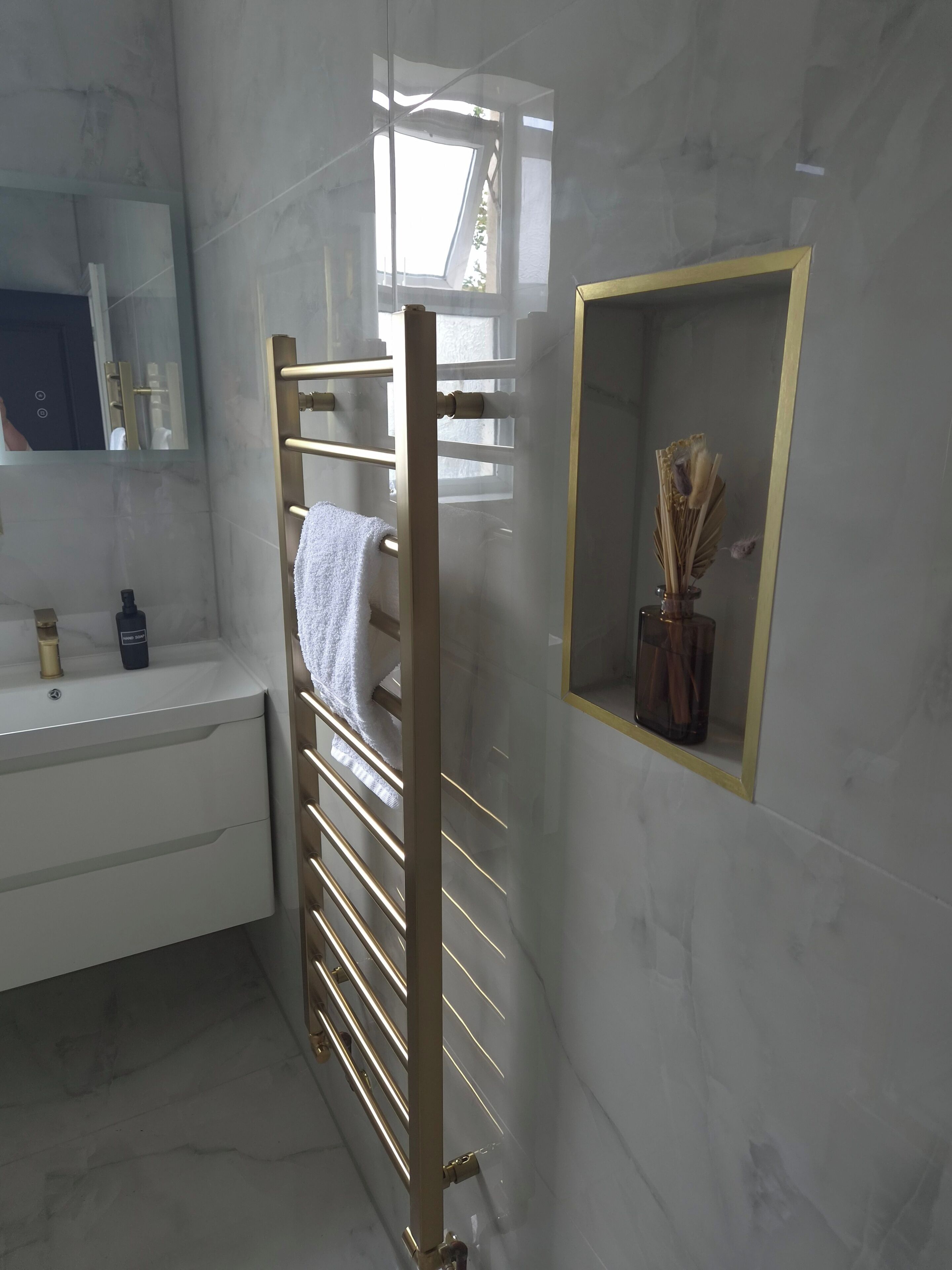 A bathroom with a white marble wall, a gold towel rack, a mirror, a sink with toiletries, and a decorative vase.