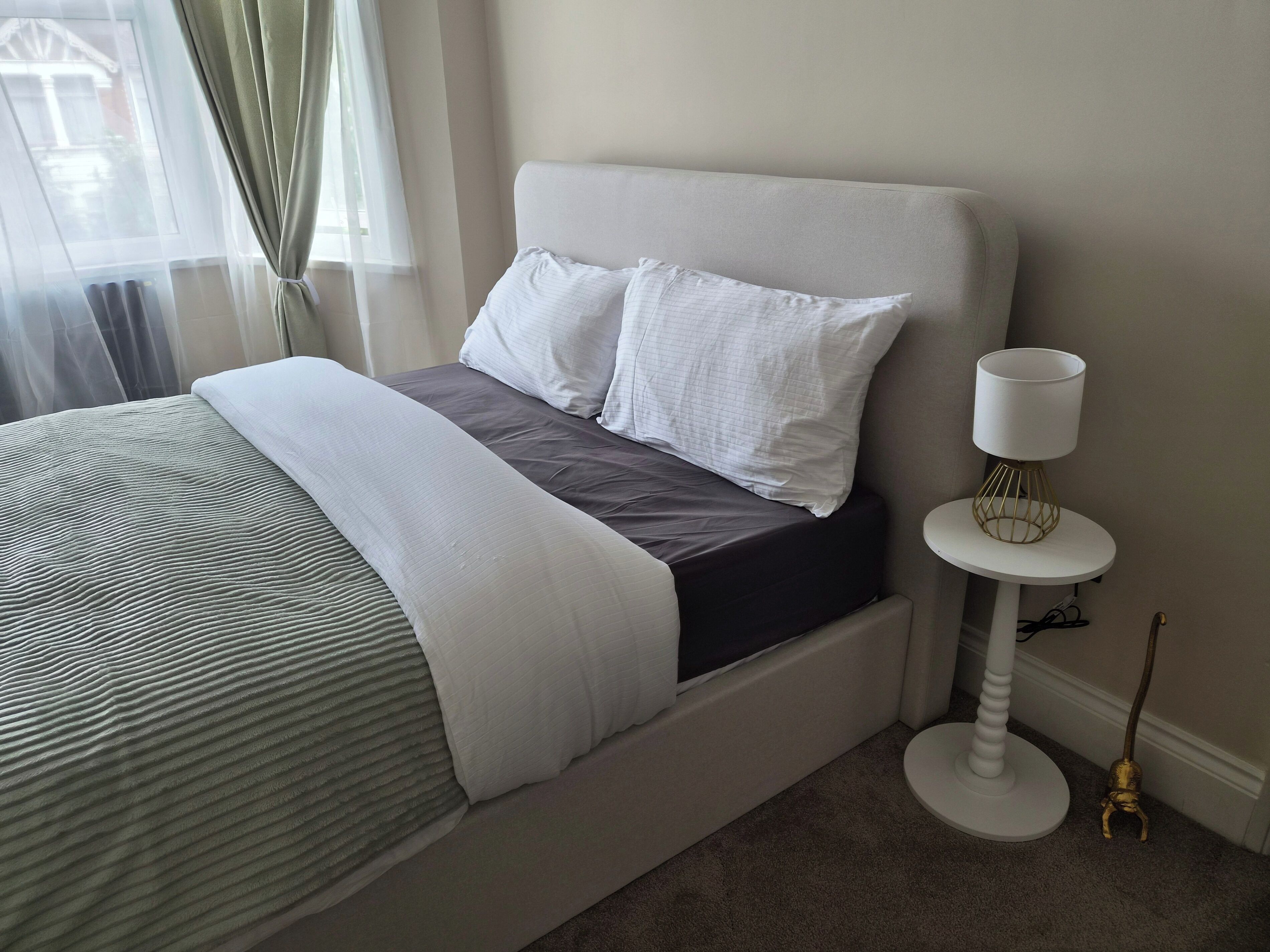 Una cama bien hecha con edredón gris y blanco, una lámpara de noche y una mesita redonda con lámpara.