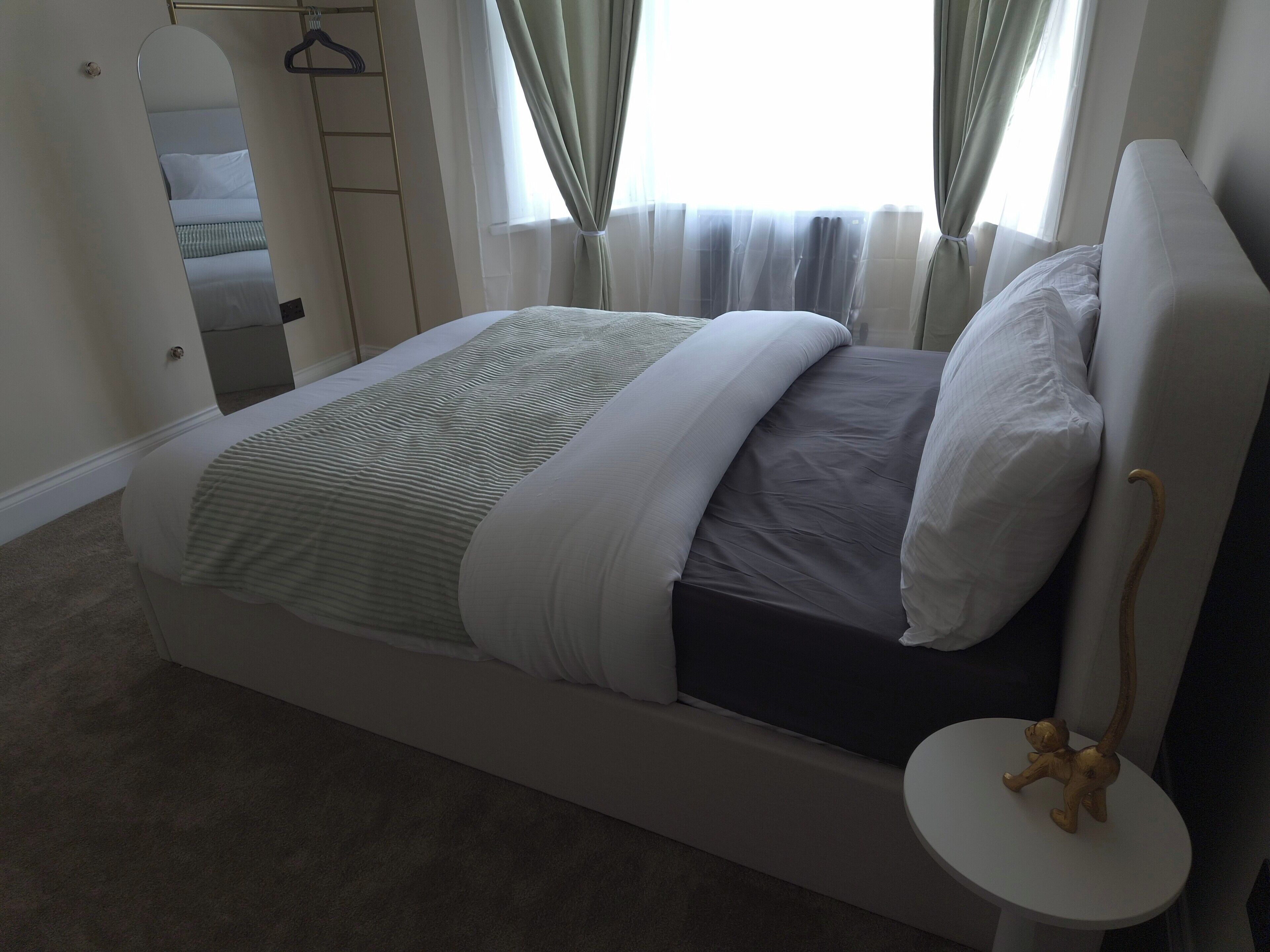 A neatly made bed with a headboard, a bedside table with a small figurine, and a window with curtains.
