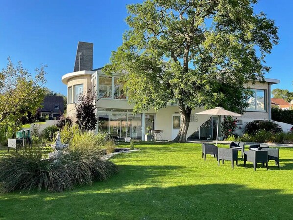 A modern house with a curved facade, a large tree in the yard, and outdoor furniture.