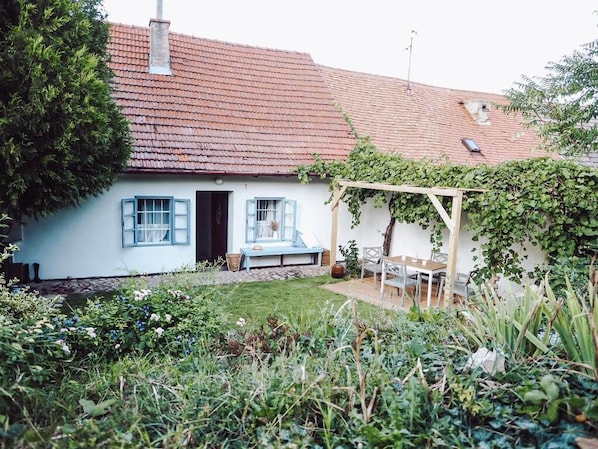 Una casa con techo de tejas rojas, fachada blanca y un jardín con banco y mesa.