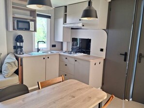 A compact kitchen with a sink, stove, and microwave. There is a dining table with chairs and a bench with a pillow. - Charming mobile home, terrace - 6 guests (Rocamadour)