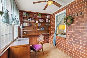 Ein Zimmer mit Schreibtisch aus Holz, einem violetten Sessel, einem Bücherregal, einem Deckenventilator und einer Backsteinwand.