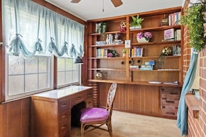 Ein Zimmer mit einem Holztisch, einem violetten Sessel, einem Bücherregal und einem Fenster mit durchsichtigen Vorhängen.