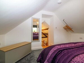 A bedroom with a sloped ceiling, a wooden bench, a purple bedspread, and a hanging clothes rack. - Ty Loti (Caernarfon)