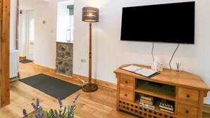 A room with a wooden floor, a black TV mounted on the wall, a wooden console table with books, a floor lamp, and a stone accent wall. - Ty Loti (Caernarfon)