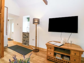 A room with a wooden floor, a black TV mounted on the wall, a wooden console table with books, a floor lamp, and a stone accent wall. - Ty Loti (Caernarfon)
