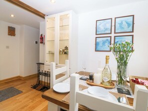 A dining area with a wooden table set for two, a bottle of Prosecco, a vase of flowers, and framed artwork on the wall. - Ty Loti (Caernarfon)