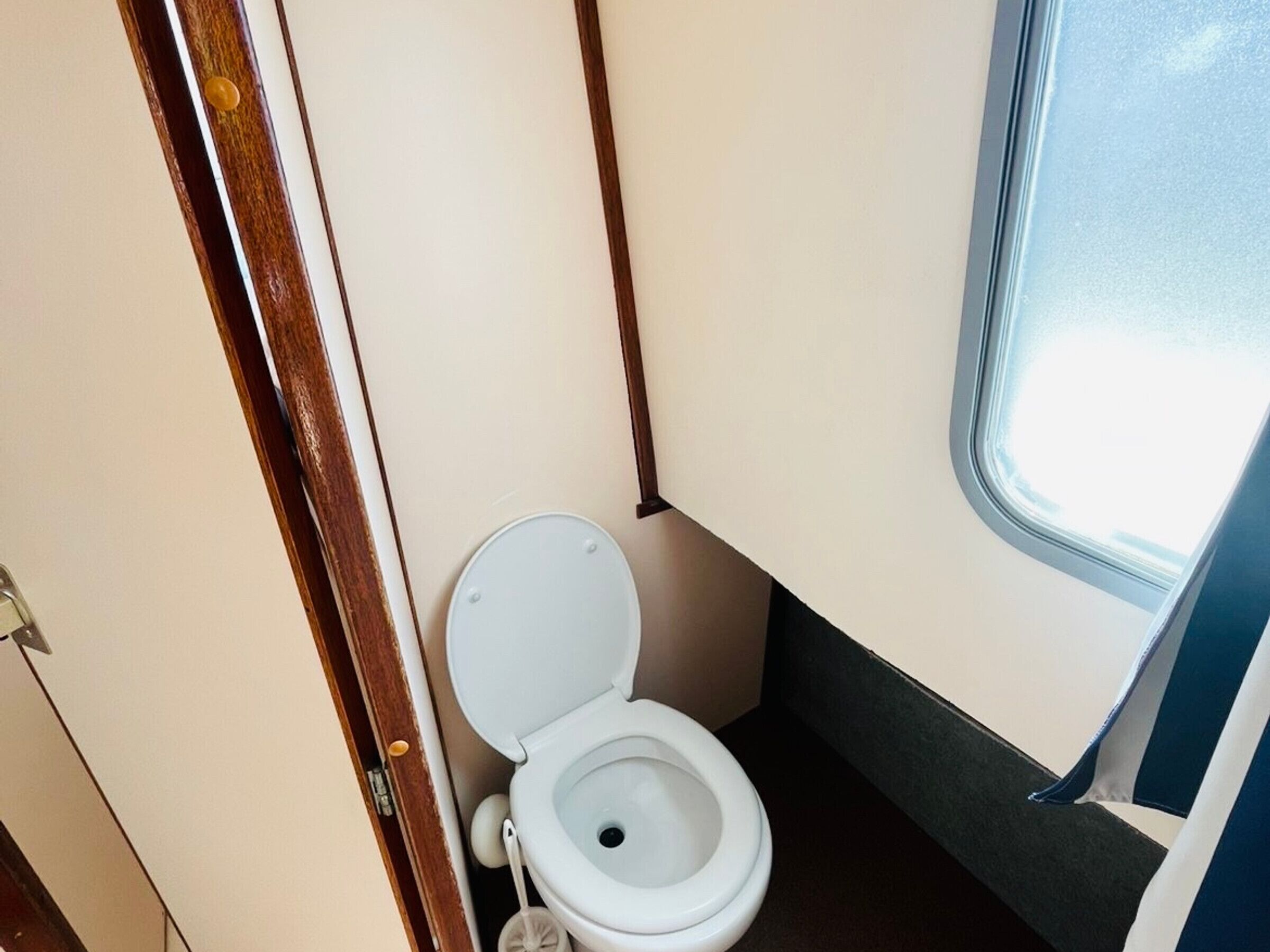 Un baño pequeño y compacto con inodoro blanco, puerta de madera y ventana que deja entrar luz natural.