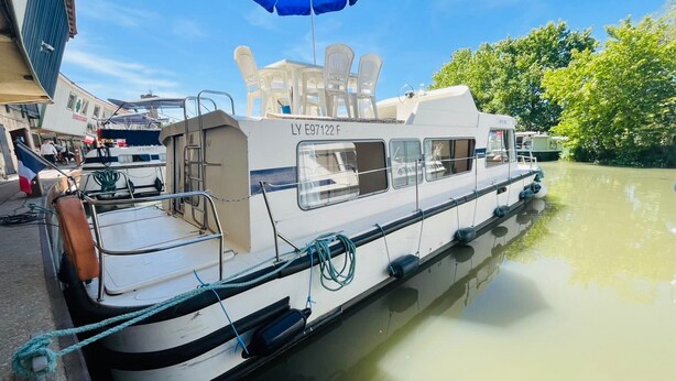 Un bateau à quai, amarré dans une marina, était équipé d’un parasol bleu, de chaises blanches et d’une structure de cabine bien visible.