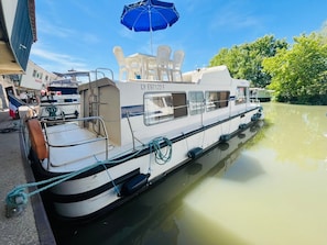 Un bote amarrado en un puerto deportivo, con un paraguas azul, sillas blancas y una estructura de cabina visible.