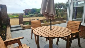 A patio with a wooden table and chairs, an umbrella, and a view of a garden and a building in the background. - Beach Horizon - ground floor flat with patio boasting views to sea (Pevensey Bay)