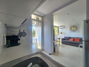 Eine moderne Küche mit Einbauofen, ein Essbereich mit Tisch und Stühlen, ein graues Sofa mit orangefarbenen Kissen und ein großes Fenster für natürliches Licht.