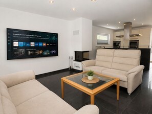 Ein modernes Wohnzimmer mit einem großen Flachbildfernseher an der Wand, der eine Streaming-Dienstanwendung anzeigt. Es gibt ein beiges Sofa und einen Glastisch mit einer kleinen Pflanze.