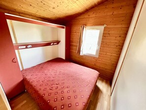 A small, single-bed room with a wooden ceiling and walls, a window with curtains, and a red bedspread.