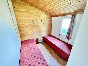 Pieni, yhden huoneen mökki, jossa on puinen katto, kaksi sänkyä punaisilla vuodevaatteilla, ikkuna verhoilla ja valkoinen ovi.