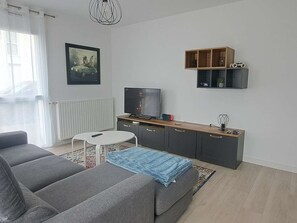 Ein Wohnzimmer mit grauem Sofa, einem Fernseher, einem kleinen weißen Tisch, einem Holzschrank und einer Heizung.