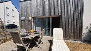 A patio with a table and chairs, a lounge chair, and a building with a wooden facade and a sliding glass door. - Comfortable apartment with parking - 4/5 guests (Carnac)