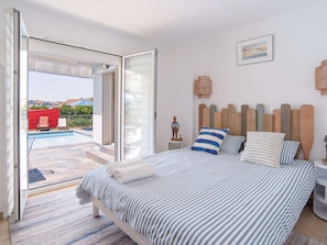 Ein Schlafzimmer mit Bett, Nachttisch, Wandlampe, einem Gemälde und Blick auf einen Außenpool.