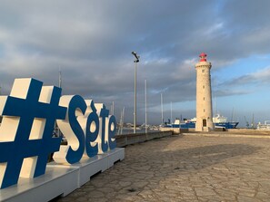 A stone-paved promenade with a large illuminated sign reading "Sete", a lighthouse, and a blue ship docked nearby. - Apartment with AC, parking - 4 guests (Sète)