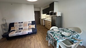 Een compact woongedeelte met een bank, een eettafel met stoelen, een keukenhoek met een magnetron en koelkast, en een plafondlamp.