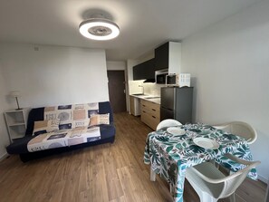 A compact living space with a sofa, a dining table with chairs, a kitchen area with a microwave and refrigerator, and a ceiling light fixture. - Studio near the beach - 4 guests (Gruissan)