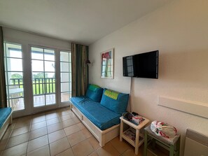 Ein Zimmer mit einer Couch, einem an der Wand montierten Fernseher, einem kleinen Tisch mit Gegenständen darauf und einer Glastür, die zu einem Außenbereich führt.