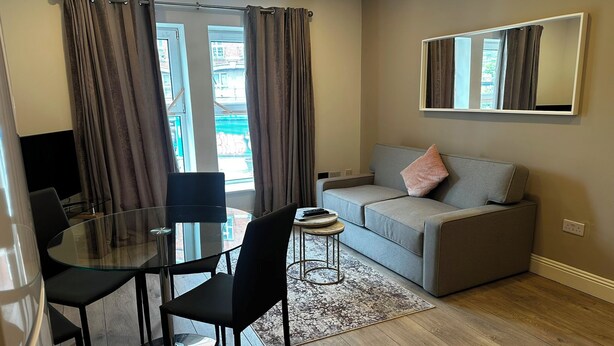 En moderne stue med et glasbord, to sorte stole, en grå sofa med en lyserød pude og et stort spejl på væggen.