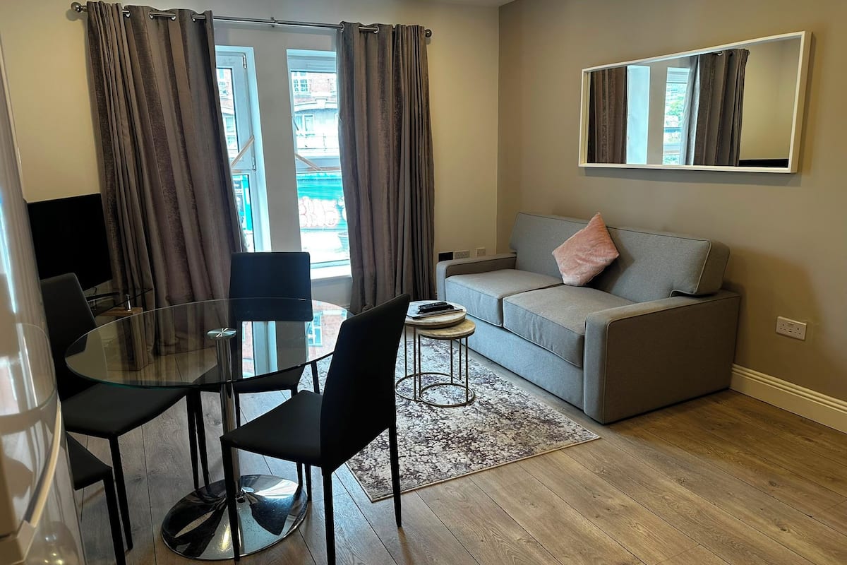 Uma sala de estar moderna com uma mesa de jantar de vidro, duas cadeiras pretas, um sofá cinza com almofada rosa e um espelho grande na parede.