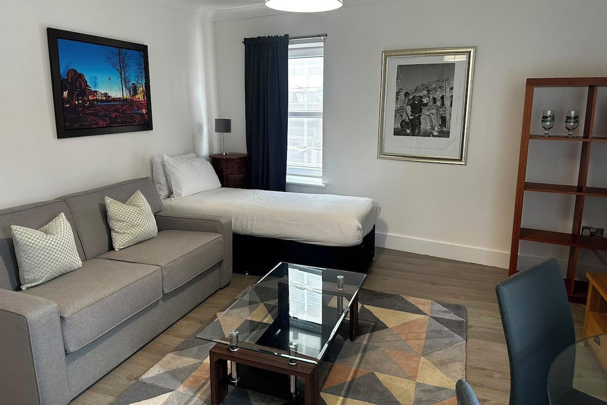 Quarto de hotel moderno com cama, sofá, mesa de centro de vidro e estante.