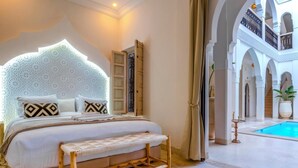 A bedroom with a large bed, a bench, a decorative headboard, and a view of a pool and courtyard. - Riad Dar Noa Marrakech (Marrakech)