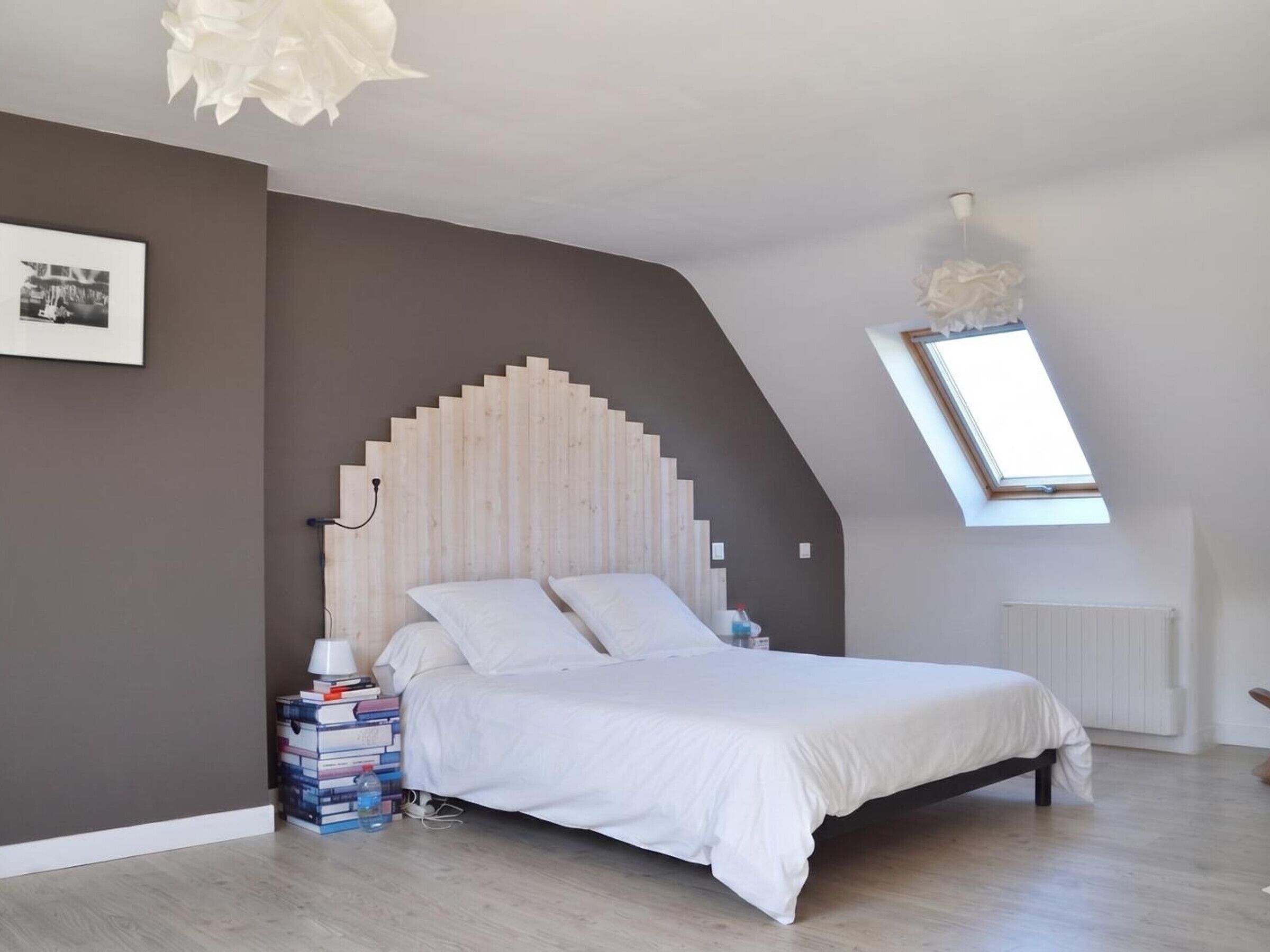 A bedroom with a sloped ceiling, a wooden headboard, a skylight, a bed, a bedside table with books, and a radiator.